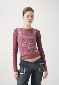 Grafisk tanktop i mauve med hvit tekst, lagd over en langermet bordeaux skjorte. Parret med mørke denim bukser med sølvdetaljer.