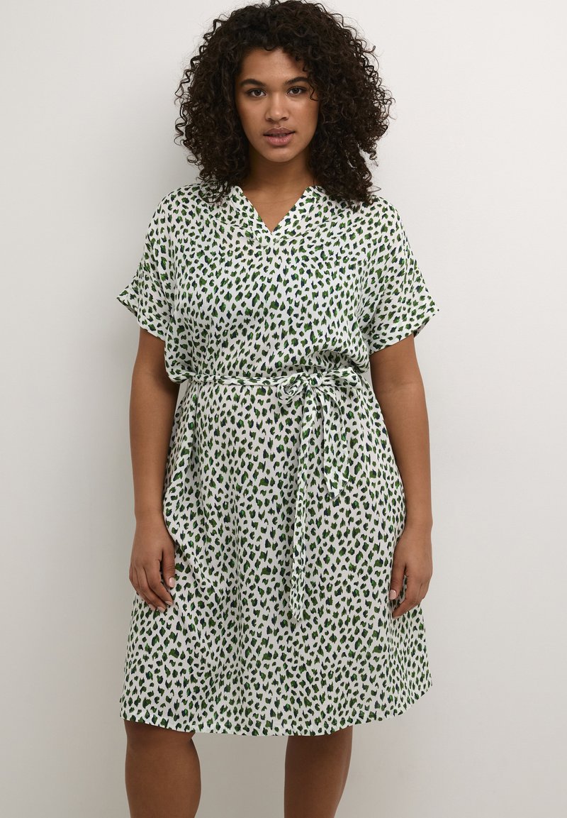 Vestido de manga corta con escote en V, base blanca y patrones abstractos en verde. Presenta una cintura anudada y una silueta fluida. Tejido suave.
