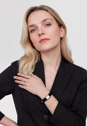 Szőke nő fekete blézerben, gyöngy fülbevalót, nyakláncot, karkötőt, ezüst gyűrűket és fekete pántos, téglalap alakú karórát visel.