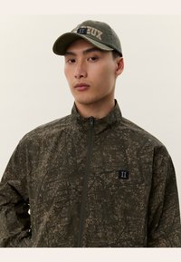 Young man wearing a dark green patterned zip-up jacket and matching cap with embroidered lettering, facing forward with a neutral expression.