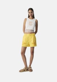 Witte mesh tanktop met "97 ATHLETIC CLUB" in goud, gecombineerd met gele op maat gemaakte shorts met borduurwerk en een riem. Bruine sandalen.