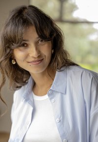 Jeune femme aux cheveux bruns ondulés, portant des boucles d'oreilles en or, un haut blanc et une chemise bleu clair à boutons, souriant à l'intérieur près d'une fenêtre.