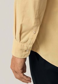 Camisa de manga larga con un tejido texturizado de color amarillo claro. Presenta un puño abotonado y un acabado suave, combinada con pantalones de color oscuro.