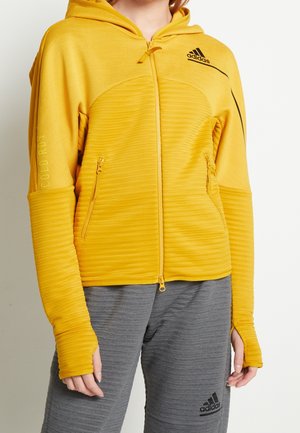 Personne portant un sweat à capuche zippé jaune Adidas avec des trous pour les pouces et un pantalon gris Adidas, debout devant un fond clair uni.