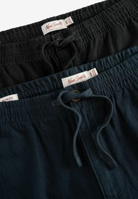 Două perechi de pantaloni expuse. Una este neagră, iar cealaltă este verde închis, ambele având talie elastică și șnururi de strângere. Etichetele sunt vizibile.