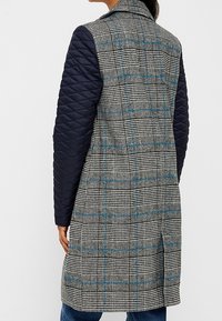 Persona che indossa un cappotto a quadri lungo fino al ginocchio con maniche in nylon blu, in piedi con la schiena rivolta verso la camera su uno sfondo semplice.
