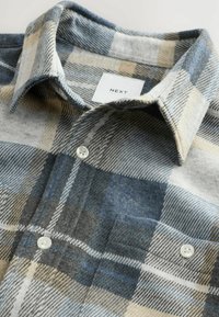 Close-up of a folded plaid flannel shirt in blue, gray, beige, and white with white buttons and a front chest pocket.