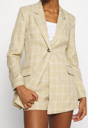Beige ternet blazer med matchende skræddersyede shorts, båret med en hvid top, vist fra midt på låret til halsen, højre hånd griber blazerens kant.