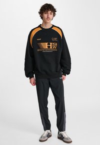 Svart sweatshirt med orange detaljer och logotyp. Kombineras med svarta byxor med grå sidopaneler och grå sneakers med blå ränder.