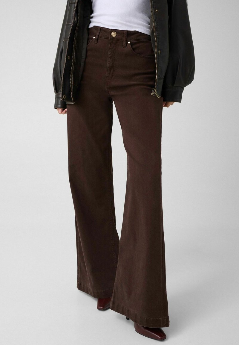 Personne portant un pantalon large taille haute marron foncé, un haut blanc, une veste noire et des chaussures pointues marron foncé sur un fond uni.