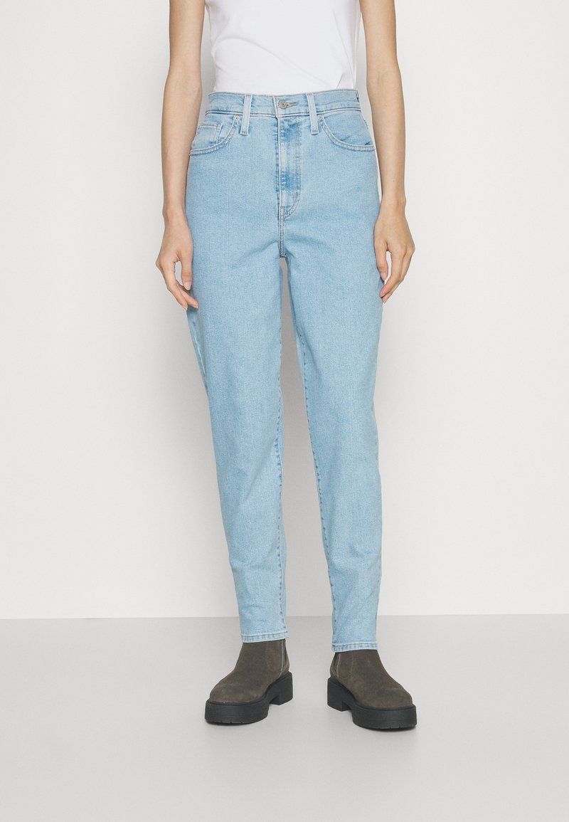 Personne portant un jean taille haute bleu clair à jambes droites, associé à des bottes épaisses foncées et un haut blanc sans manches.