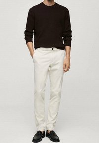 Weiße Chinos kombiniert mit einem dunkelbraunen gerippten Pullover und schwarzen Slippern. Die Hose hat eine flache Vorderseite und einen einzelnen Knopfverschluss.
