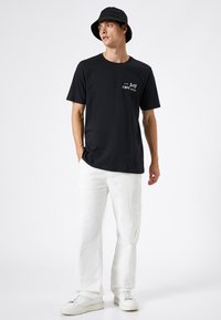 Czarny t-shirt z krótkim rękawem z napisem "Self care", w połączeniu z białymi spodniami cargo i czarnym kapeluszem typu bucket, na jednolitym tle.