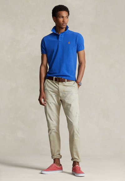 Μπλε πόλο μπλουζάκι με ριμπ γιακά, μπεζ παντελόνι με πιτσιλιές μπογιάς και ροζ αθλητικά παπούτσια. Το μοντέλο φοράει καφέ ζώνη και ρολόι.