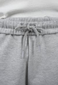 Gros plan sur la taille d’un pantalon de jogging gris clair avec un cordon noué et une bande élastique, porté avec un tissu blanc visible au-dessus.