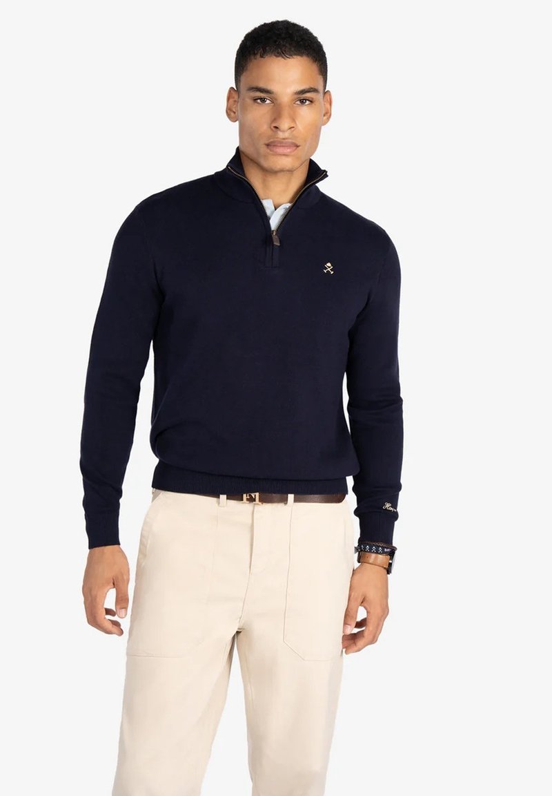 Suéter de punto azul marino con cuello con cremallera de un cuarto, que presenta un pequeño logotipo en el lado izquierdo del pecho. Combinado con pantalones beige claros.