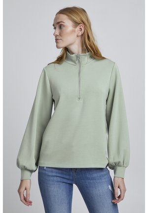 Mintgrüner Pullover mit halbem Reißverschlusskragen, langen Puffärmeln und einer weichen Textur, kombiniert mit hellblauen, distressed Jeans.