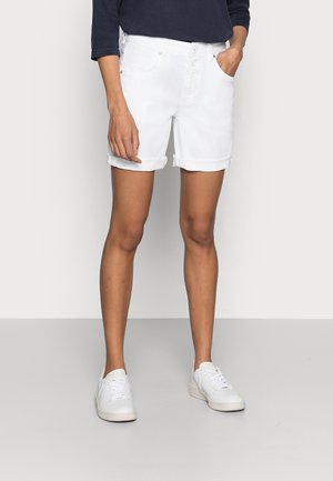 Person iført hvide shorts med opsmøg, hvide sneakers og en mørk langærmet top, stående med den ene hånd i lommen mod en hvid baggrund.