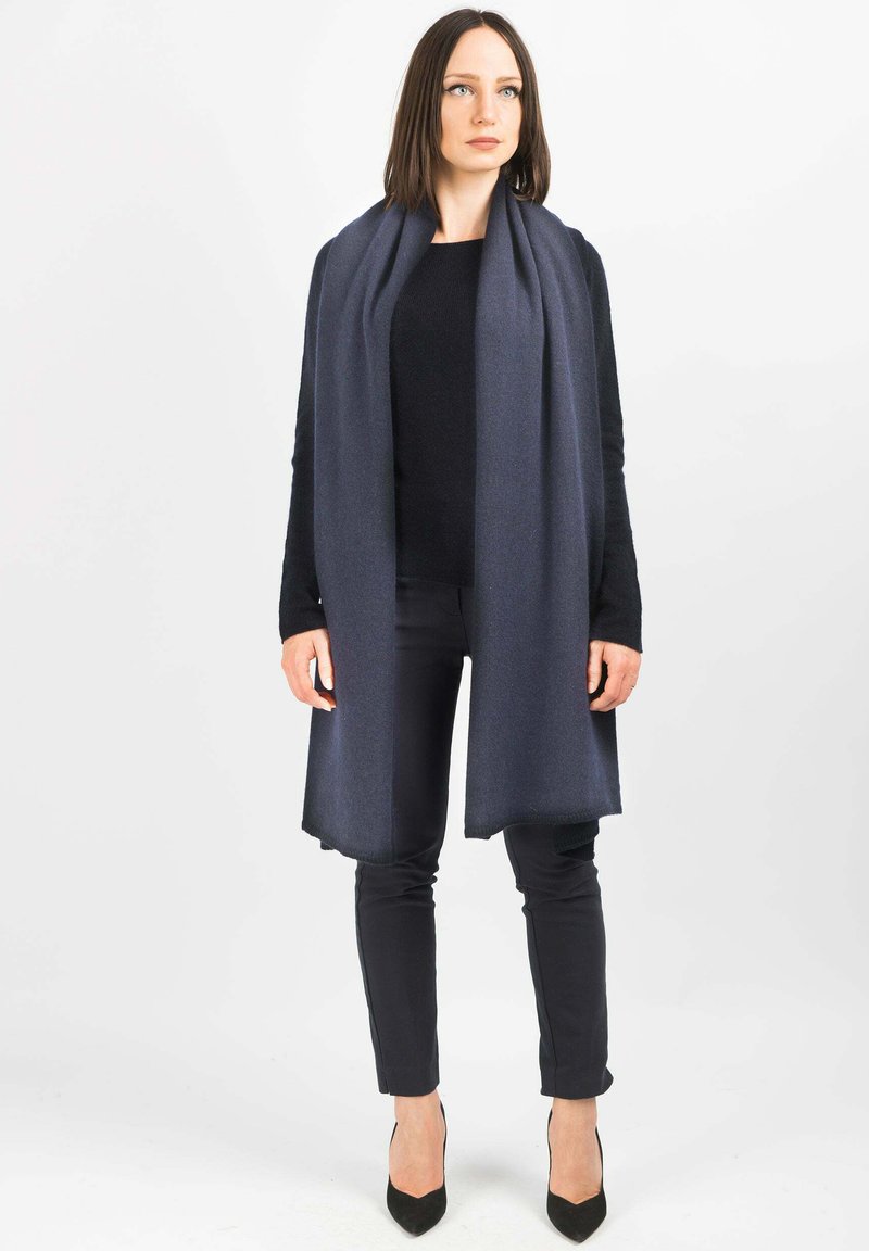 Sciarpa di lana blu scuro drappeggiata sulle spalle, con una texture liscia e bordi frangiati. Indossata con un top nero e pantaloni slim fit.