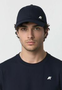 Jeune homme à la peau claire et aux yeux verts, portant une casquette bleu marine et une chemise bleu marine, toutes deux ornées d'un logo stylisé "K" blanc.