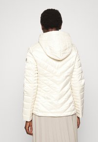 Kremowa kurtka puchowa z kapturem, z haftem w kształcie zygzaka oraz gładką, błyszczącą teksturą. Lekka z dopasowanymi rękawami i prostym dołem.