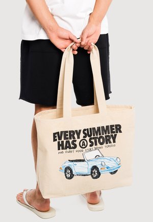 Osoba w czarnych szortach i białych klapkach trzyma beżową torbę z niebieskim rysunkiem samochodu i napisem "Every summer has a story" na jasnym tle.