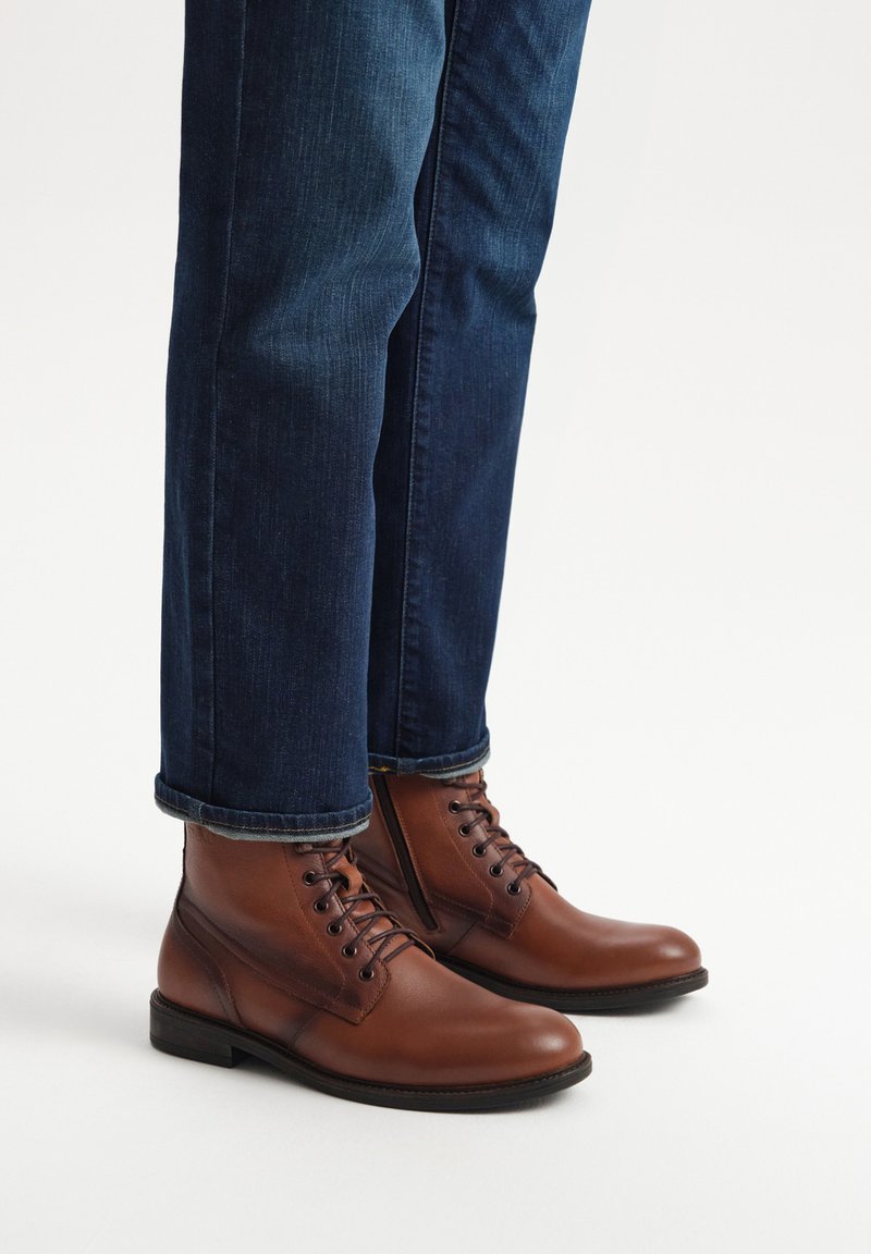 Brązowe skórzane botki za kostkę z sznurowanym wzorem i mocnymi podeszwami, noszone z ciemnoniebieskimi dżinsami, z podwiniętymi mankietami.