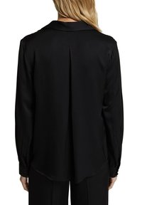 Femme aux cheveux blonds portant un chemisier noir à manches longues avec un pli au centre du dos, associé à un pantalon noir.