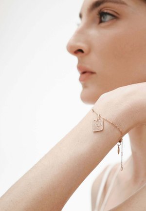 Delicada pulsera de oro rosa con una etiqueta cuadrada y eslabones de cadena, que presenta un pequeño charm. Superficie lisa y detalles intrincados visibles.