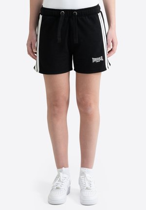 Persona que lleva puestos pantalones cortos negros con rayas laterales blancas y el logo "Lonsdale London", calcetines blancos y zapatillas blancas, de pie frente a un fondo blanco.