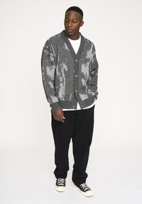 Grå stickad cardigan med ett strukturerat mönster, V-ringad och knappöppning, bärs över en vit skjorta. Matchas med svarta byxor och sneakers.