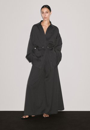Modèle portant une chemise noire à rayures et un pantalon large, les mains dans les poches, se tenant devant un fond neutre.
