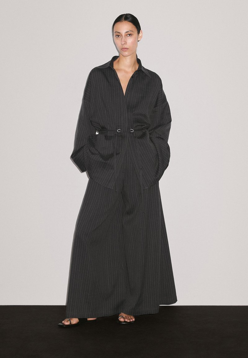 Model wearing a black pinstripe oversized shirt and wide-leg pants, with hands in pockets, standing against a neutral background.