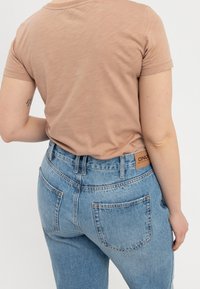 Svetle hnědé tričko s krátkým rukávem, přiléhavé; vysoce posedlé modré džíny s vybledlým povrchem a viditelnou značkou na zadním pásu.