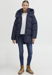 Navy puffer jacket with a high collar and zip closure, featuring a quilted design. Paired with denim and tan boots with laces.