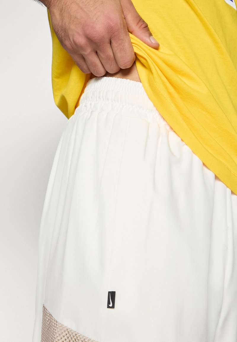 Weiße Sportshorts mit elastischem Bündchen, versehen mit Mesh-Akzenten an den Seiten und einem kleinen schwarzen Nike-Logo-Tag. Ein knallgelbes Shirt bedeckt teilweise die Shorts.