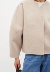 Smėlio spalvos vilnos mišinio perverstas švarkas su ilgomis rankovėmis, laisvu siluetu ir struktūrizuota apdaila. Dėvimas ant balto marškinėlio, derinamas su mėlynomis džinsomis.