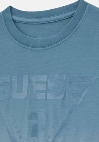 Camiseta de algodón azul con cuello redondo, que presenta un logotipo gráfico en relieve en un tono más claro, con la palabra "GUESS" y otros elementos textuales.