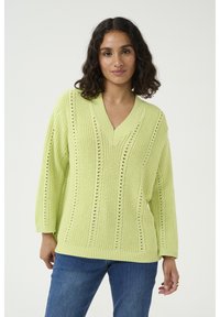 Pull en maille surdimensionné vert citron avec un col en V, orné de rayures verticales et de détails ajourés, associé à un jean bleu.