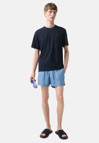 Marine kortærmet t-shirt, lyseblå badebukser og marine slidesandaler. Modellen holder solbriller med lilla linser og står foran en neutral baggrund.