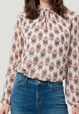 Frau trägt eine weiße Langarmbluse mit floralem Muster, die in eine blaue, hoch taillierte Jeans gesteckt ist.