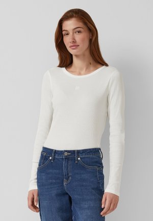 Pikkade punaste juustega naine, kes kannab valget pikkade varrukatega vormiriietust ja siniseid teksaseid, seisab ühetaustalise halli tausta ees.