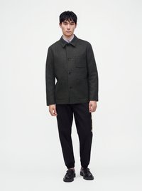 Un homme se tient debout portant une veste à carreaux foncée, un pantalon noir et des chaussures noires sur un fond clair uni.
