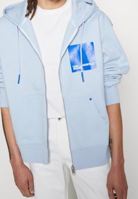 Felpa con cappuccio azzurro chiaro con zip, cappuccio con coulisse, tasche frontali e una toppa grafica blu con la scritta "Karl Lagerfeld Jeans". Tessuto dalla consistenza morbida.