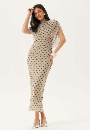 Kvinna som ler, klädd i en beige figurnära klänning med bruna prickar och ett genomskinligt matchande tyg som är draperat över axlarna, silverfärgade klackar, vitt armband.