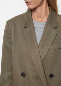 Dvojtečný blazer v olivově zelené tkanině, s klopovým límcem, dvěma černými knoflíky a přední kapsou, vrstvený přes šedý top.
