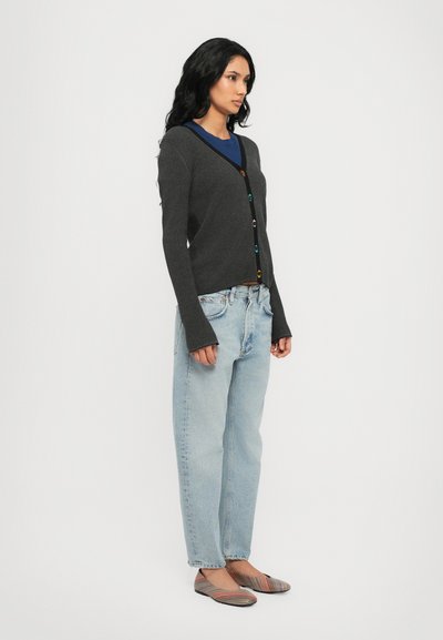 Cardigahn grigio a coste con bottoni multicolori, maniche lunghe e scollatura a V, indossato sopra una maglietta blu, abbinato a jeans blu chiaro e ballerine a righe.