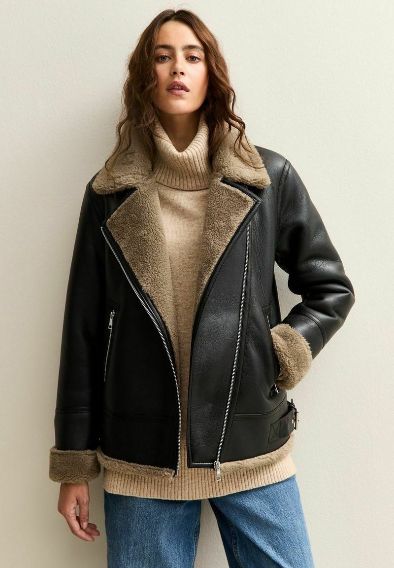 Donna che indossa un giubbotto aviator in pelle nera con rivestimento in shearling beige, un maglione beige a collo alto e jeans blu, in piedi contro un muro crema.