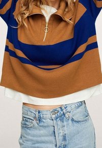 Pull en rayures marron et marine avec un col zippé quart, doté d'un tirette en métal rond, porté par-dessus une chemise blanche et un jean bleu clair.