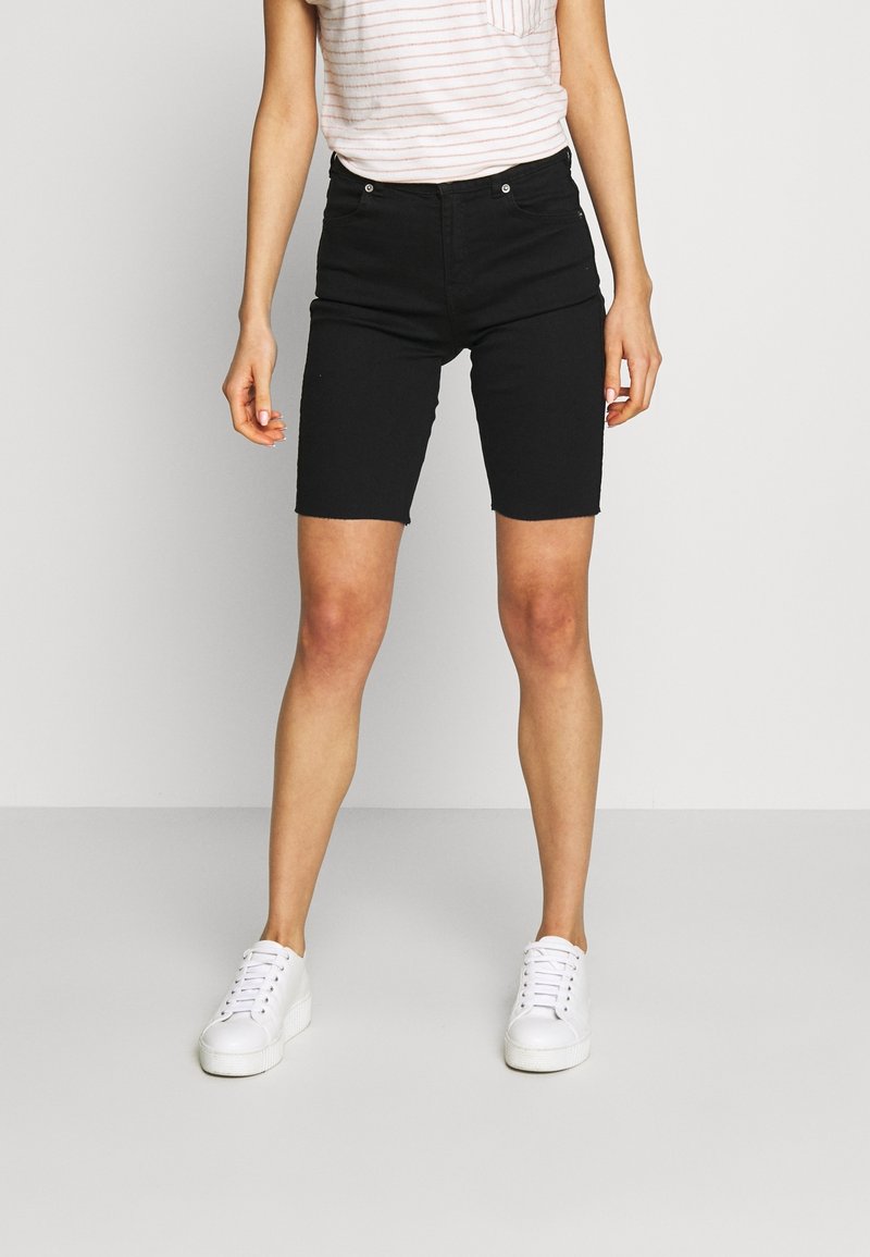 Personne portant un short noir au genou, un t-shirt blanc rayé de rose et des baskets blanches, debout devant un fond uni.